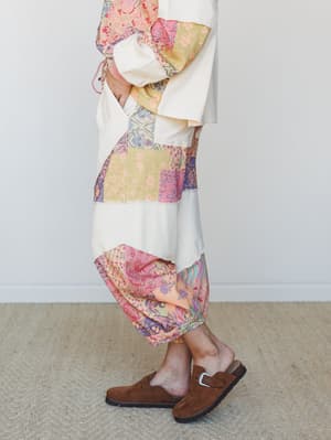 A side-profile shot of a patchwork midi-length pant in soft cream and mixed floral prints, styled with brown suede clogs for an effortlessly earthy, lived-in look.