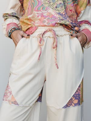 A mid-shot focused on cream wide-leg drawstring pants with patchwork floral print insets at the hem, styled with a matching mixed-print top and layered boho jewelry — the kind of outfit that feels intentionally relaxed without trying too hard.