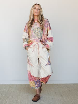 A full-body model shot of a dreamy mixed-print set — an oversized patchwork top layered over cream wide-leg parachute pants with a pink drawstring, styled with brown suede clogs and layered jewelry for an effortlessly boho, lived-in feel.