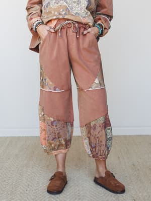 A mid-shot of relaxed patchwork jogger pants in a worn mocha tone, pieced together with floral tapestry fabric panels at the lower leg and styled with beaded bracelets, a floral sweatshirt, and suede clogs — the kind of outfit that looks effortless b