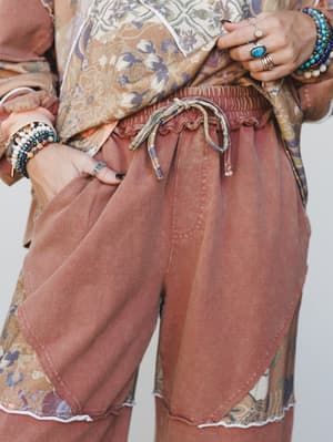 A close-up torso shot spotlighting relaxed drawstring pants in washed mocha layered over a floral patchwork hem, styled with an abundance of stacked rings and beaded bracelets that give the whole look an effortlessly collected, boho-market feel.