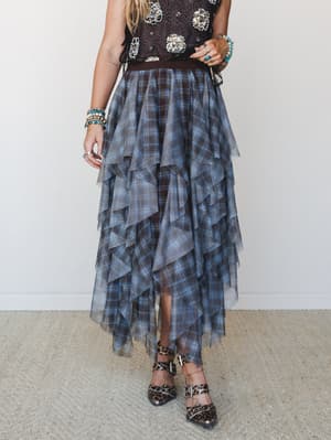 A tiered plaid maxi skirt with a sheer tulle overlay cascades in dramatic ruffled layers, styled with an embellished dark top, leopard print heels, and a stack of turquoise and mixed bead bracelets — the kind of outfit that makes a statement without