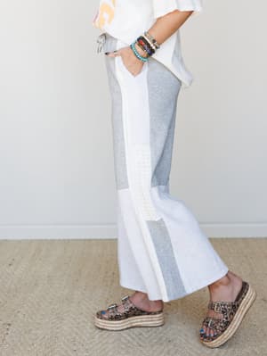 A side-angle shot of wide-leg pants in heather grey and white colorblock with a crochet lace inset panel down the leg, styled with a white graphic tee, stacked beaded bracelets, and leopard print platform sandals — relaxed and put-together at the sam