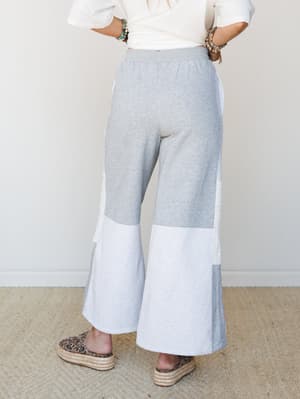 Back view of wide-leg colorblock sweatpants in heather grey and light grey with lace side panel detailing, styled with a knotted white tee, stacked boho bracelets, and platform espadrilles — comfy-cool with a little personality.