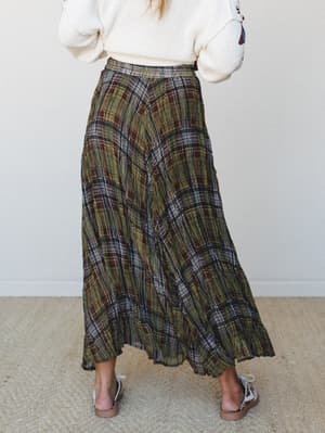 Shot from behind, a flowing plaid maxi skirt in earthy olive and brown tones is styled with a cream chunky knit sweater — the kind of autumn outfit that makes you want to walk through leaves and pretend you have nowhere to be.