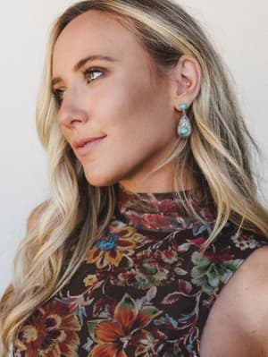 A close-up portrait of a model wearing turquoise and silver teardrop earrings, paired with a rich dark floral top — the kind of shot that sells the accessory by making you feel the whole vibe of the outfit.