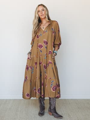A flowy tiered maxi dress in warm camel with bold floral print, styled with dark cowboy boots and layered jewelry — the kind of outfit that feels effortless but clearly intentional.