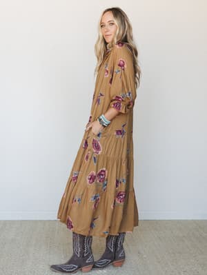 A flowy camel floral maxi dress with a tiered skirt is styled with dark cowboy boots and stacked turquoise bracelets — the kind of outfit that feels equally at home at a fall market or a friend's backyard wedding.