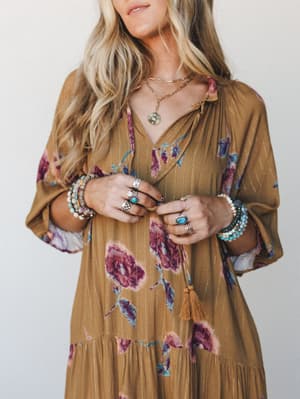 A golden camel floral dress with bell sleeves and a tassel tie neckline, styled with layered gold necklaces and stacked turquoise and silver rings and bracelets — the kind of outfit that looks like it was effortlessly assembled but very much wasn't.