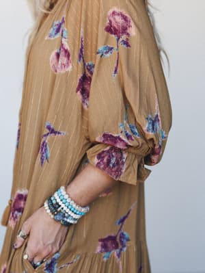A close-up detail shot of a camel-toned floral maxi dress with gold metallic pinstripes and puff sleeves, styled with a stack of beaded bracelets and silver rings — earthy and expressive in equal measure.