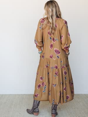 A flowing camel floral maxi dress with a tiered skirt and balloon sleeves, styled with dark cowboy boots and stacked bracelets — shot from the back against a clean white wall, giving it a relaxed, lived-in boho feel.