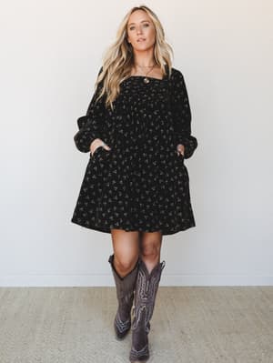 A flowy black mini dress with a delicate ditsy floral print, square neckline, and long sleeves pairs effortlessly with tall gray cowboy boots — the kind of outfit that feels equally at home at a weekend market or a casual night out.