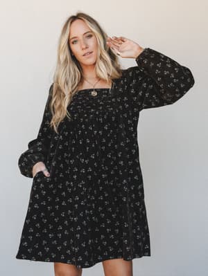 A flowy black mini dress with a ditsy floral print and long bishop sleeves, styled with layered gold necklaces against a soft neutral backdrop — the kind of dress that works just as hard at brunch as it does at a fall concert.