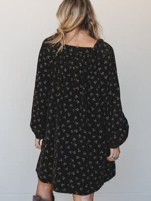 Shot from the back, a flowy black mini dress with a ditsy floral print and billowy long sleeves has that effortless, worn-it-a-hundred-times feel — styled with silver rings, a delicate gold necklace, and brown boots peeking in at the hem.
