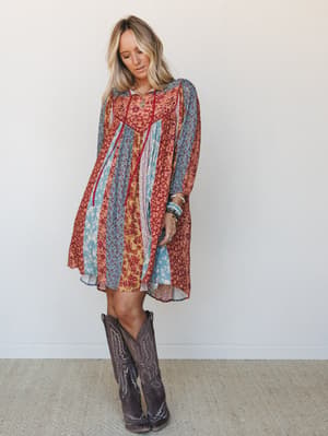 A flowy patchwork mini dress in warm rust and dusty blue florals, styled with tall brown cowboy boots and layered bracelets — the kind of outfit that looks like it wandered in from a good story.