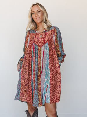A flowy patchwork mini dress in warm mixed florals — rust, teal, and mustard panels sewn together with deep red piping — styled with cowboy boots and layered gold jewelry for that effortless boho-meets-western feel.