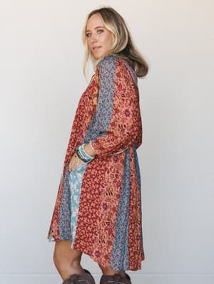 A patchwork floral midi dress in warm rust, dusty blue, and terracotta panels is styled with cowboy boots and stacked bracelets — shot from behind in a three-quarter turn that shows off the relaxed swing silhouette and lived-in boho energy.