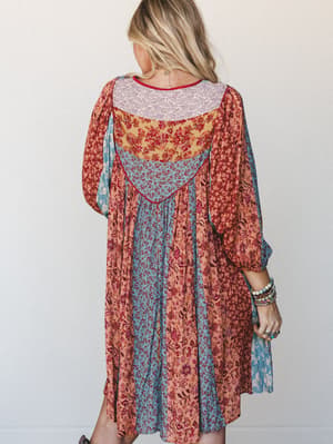Shot from the back, this flowy midi dress layers multiple mixed floral prints in a patchwork arrangement — warm terracotta and dusty blue panels meet marigold and cream, styled with stacked beaded bracelets and a delicate gold necklace for an effortl