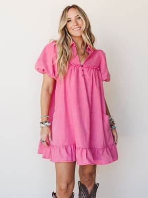 A bright pink babydoll mini dress with ruffle hem and puff sleeves, styled with brown cowboy boots and stacked turquoise bracelets against a clean studio wall — playful and warm with just enough western edge.