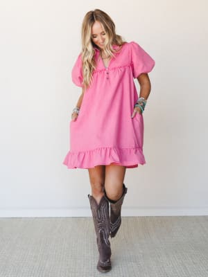 A bright orchid pink mini dress with puff sleeves and a ruffle hem gets a Western twist paired with dark brown cowboy boots and stacked turquoise bracelets — playful, warm, and full of personality.