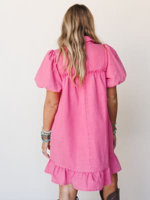 A bright orchid pink mini dress shot from the back, showing off puff sleeves, ruffle hem, and a relaxed babydoll silhouette — styled with stacked beaded bracelets and cowboy boots for that sweet spot between playful and western.
