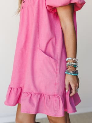 A close-cropped shot of a bright pink mini dress with a ruffle hem and side pocket, styled with a stacked beaded bracelet arm party — casual and joyful with a soft, lived-in feel.