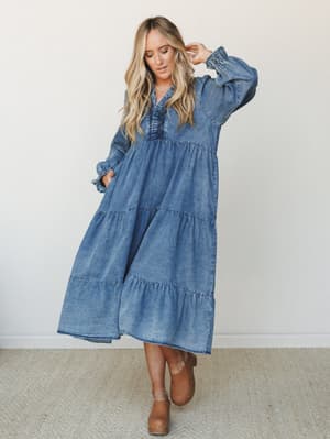 A flowy washed denim maxi dress with tiered skirt and billowy sleeves, styled with tan clogs for an effortless boho-meets-western feel — the kind of dress that looks equally at home at a farmers market or a backyard gathering.