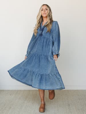 A flowy washed denim maxi dress with tiered skirt and smocked bodice, styled with tan clogs — the kind of dress that makes a Tuesday feel like a road trip.