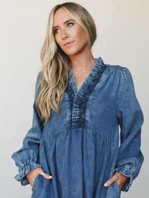 A relaxed denim maxi dress with smocked and ruffled v-neckline detailing, worn with easy hands-in-pockets confidence against a soft neutral studio backdrop — the kind of piece that feels like a whole outfit without trying.