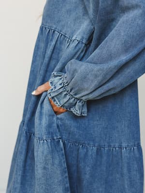 A close-up detail shot of a washed denim maxi dress, showing the tiered skirt construction and a billowy long sleeve with a gathered ruffle cuff — the kind of dress that feels like your favorite jeans but prettier.