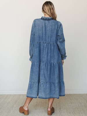 A washed denim maxi dress shot from the back, showing off its tiered skirt, ruffled neckline, and long billowy sleeves — the kind of dress that feels like a denim jacket had a very romantic moment.