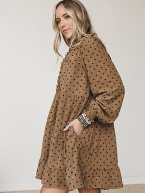 A corduroy babydoll mini dress in warm mocha with an all-over black polka dot print, styled with stacked beaded bracelets and a silver ring — the kind of dress that feels like a hug but looks like you tried.