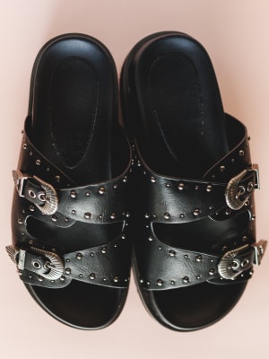 Black sandals with studded details and buckles on a pink background