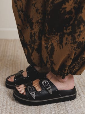 Black sandals with buckles worn by a person in patterned pants on a neutral background