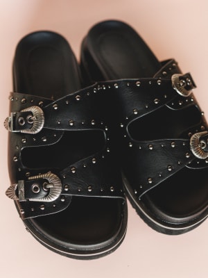 Black sandals with metallic buckles and studs on a light pink background