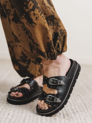 Black sandals with buckles worn with brown pants on a neutral background