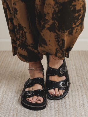 Black studded sandals worn with patterned pants on a neutral background