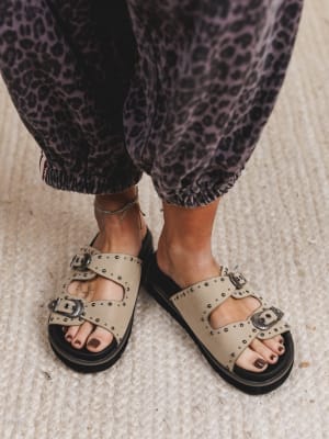 Beige sandals with black soles worn with leopard print pants on a neutral background
