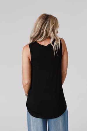 A woman with long, wavy blonde hair is shown from the back wearing The Nest Shelli Scoop Neck Tank Top in black and light blue jeans, standing against a plain light gray background.