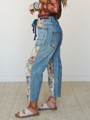 A side-profile shot of relaxed-fit denim jeans with floral tapestry side panels, styled with a rust floral mesh top, stacked bracelets, and white buckle sandals — the kind of jeans that feel like a whole personality.