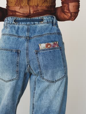 A close-up back view of medium-wash wide-leg jeans with floral patchwork peeking from the right pocket and grommet waistband detail, paired with a sheer floral lace top.