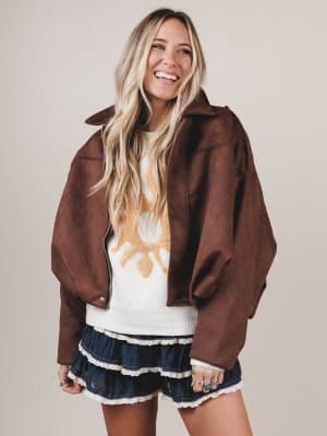 Woman wearing a brown jacket over a white sweater with a star design, standing against a plain background.