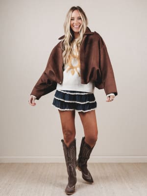 Woman wearing a brown jacket, white sweater with a graphic, blue skirt, and cowboy boots on a plain background