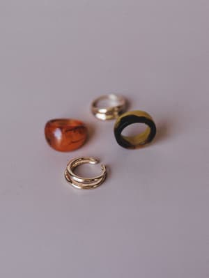Four rings scattered loosely on a soft gray surface — a translucent amber dome, a tortoise-pattern statement ring, a polished gold band, and a double-band open ring — the kind of mix that looks like it happened naturally on a nightstand.