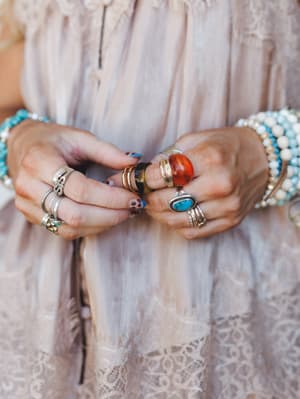 A close-up of stacked hands layered with an eclectic mix of stone rings and beaded bracelets against a blush lace dress — the kind of jewelry collection that looks like it was gathered over a lifetime of good taste.