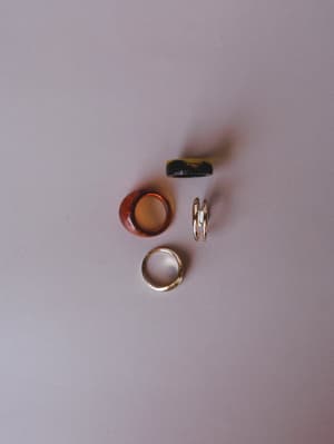 A casual flat lay of four rings grouped together — a chunky tortoise resin ring, a black-and-olive marble ring, a slim gold band, and a delicate double-band ring — all resting on a soft lavender-gray surface with an effortless, collected feel.