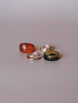 Four rings scattered casually on a soft mauve surface — an amber resin dome, a double-band gold ring, a domed gold ring, and a wide tortoise-marbled resin band that anchors the whole earthy, collected feel.