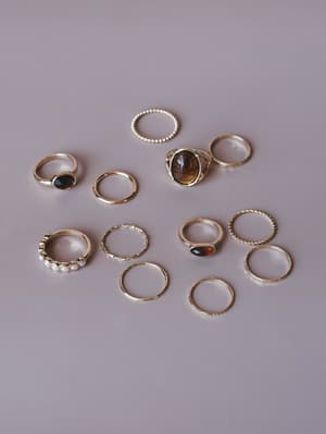 A scattered flat lay of a gold ring stack set — mixing delicate bands, pearl accents, stone statement pieces, and twisted textures against a soft mauve-grey background.