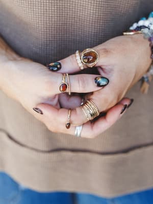 A close-up of layered hands showing off a collected stack of gold rings — stone-set, pearl-accented, twisted band, and stacked styles — worn against a taupe waffle-knit top, with a gemstone bracelet peeking in at the edge.