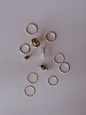 A scattered flat lay of mixed gold rings — thin bands, textured stackers, and stone-set statement pieces — arranged casually on a soft mauve surface, giving off that 'pulled from a jewelry dish' kind of energy.